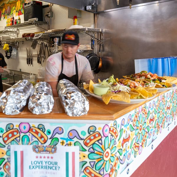 Restaurant worker prepares food behind a counter displaying foil-wrapped items and a loaded plate of nachos.