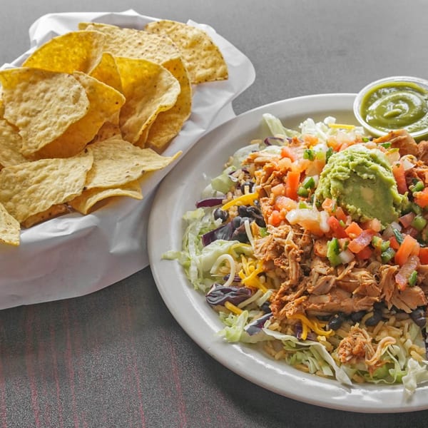 Loaded taco salad with shredded meat, beans, vegetables, guacamole, and salsa beside a basket of tortilla chips.
