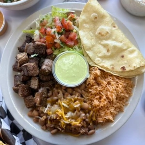 A plate of carne asada, rice, beans with cheese, lettuce, pico de gallo, tortillas, and green sauce.