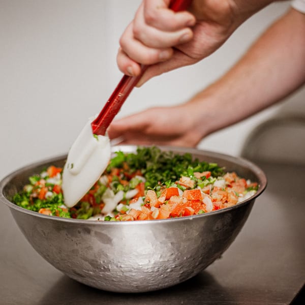 A hand mixes fresh pico de gallo with a red and white spatula in a metal bowl.