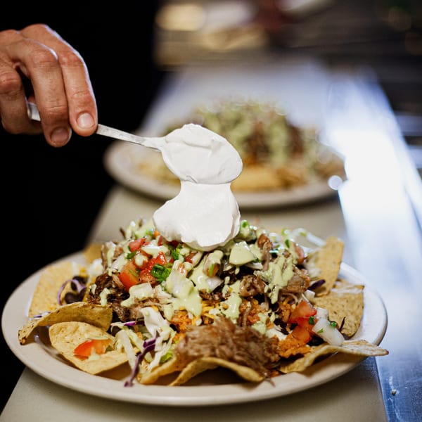 A hand spoons sour cream onto a plate of Hey Juan Burritos' loaded nachos with meat and vegetables.