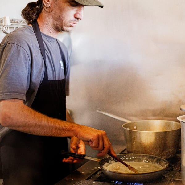A chef in a black apron and cap stirs a creamy sauce in a pan on a stove.