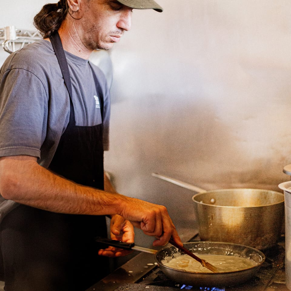 A chef in a black apron and cap stirs a creamy sauce in a pan on a stove.