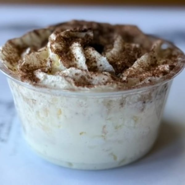 A clear plastic cup filled with a creamy dessert, topped with whipped cream and dusted with cocoa powder.